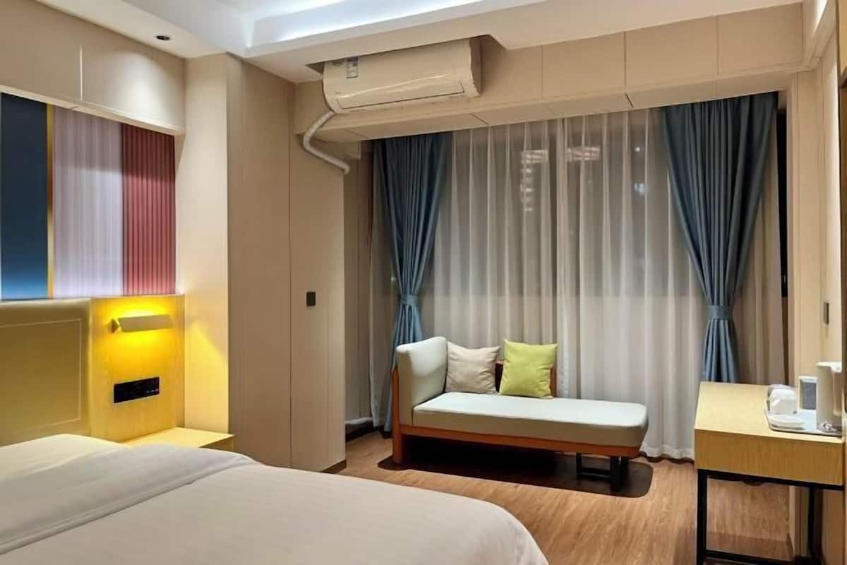 Habitación de hotel con cama, mesita de noche, sofá, ventana con cortinas y equipo de aire acondicionado.