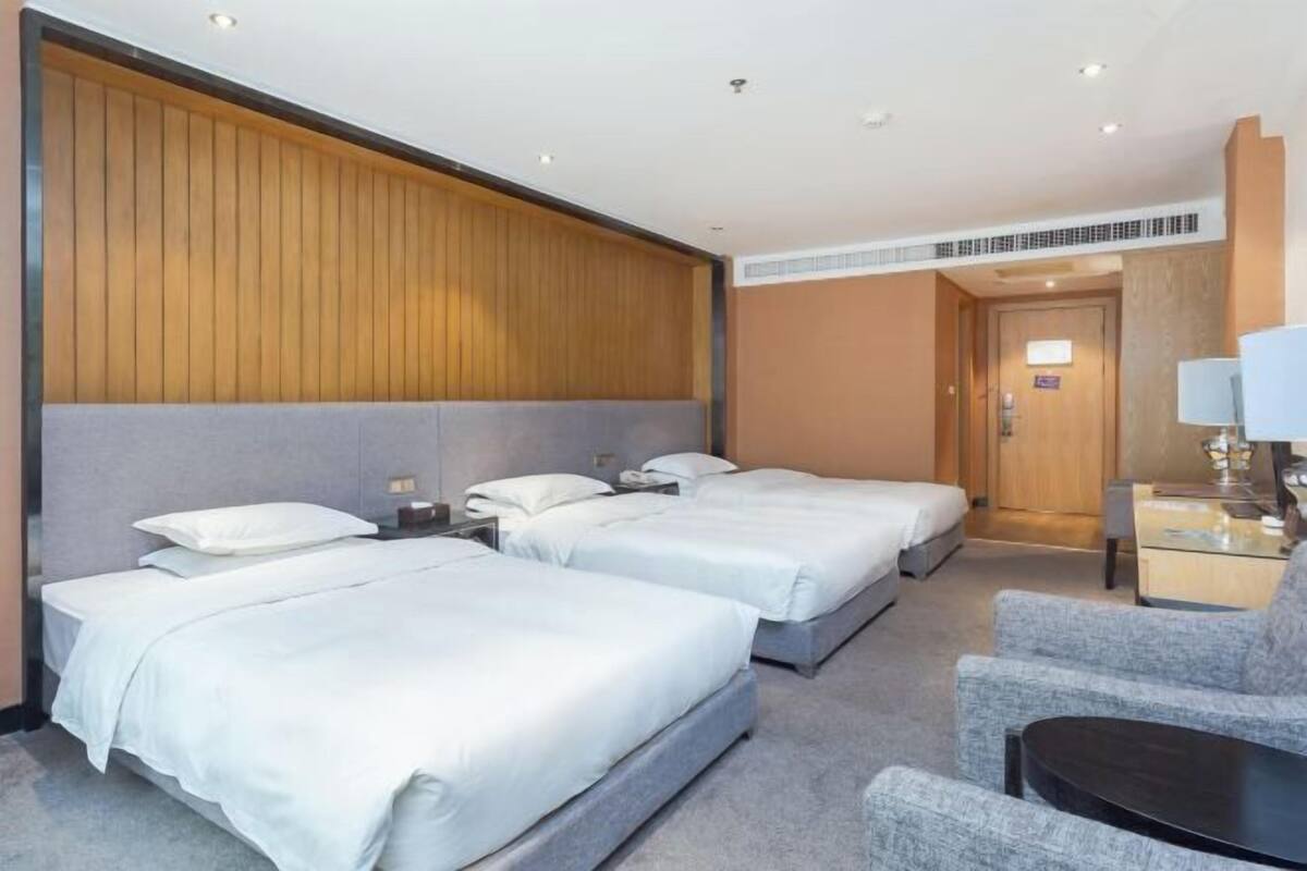 Habitación de hotel con dos camas individuales, un pequeño escritorio con lámpara y un rincón de descanso con una silla y una mesa.