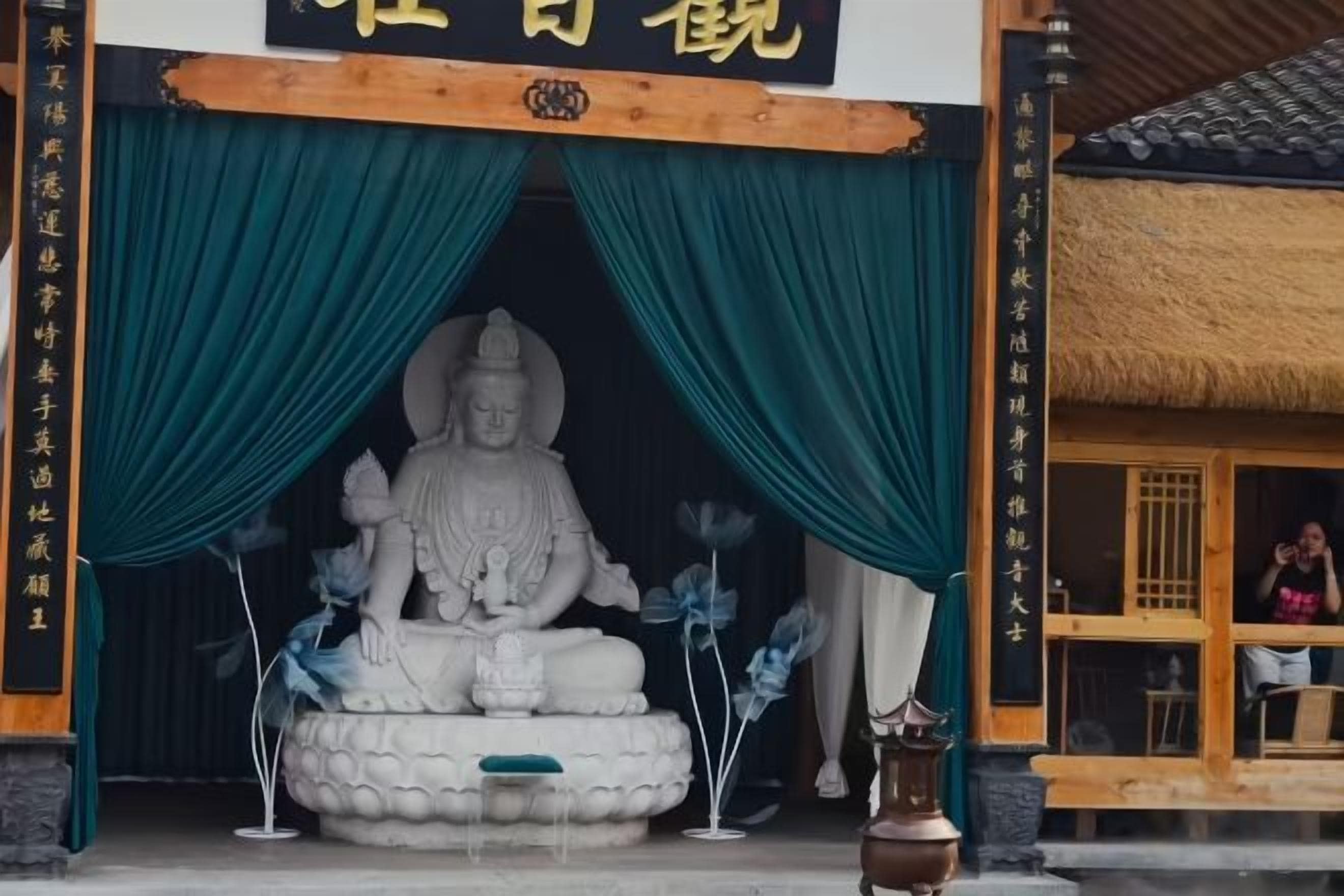 A Buddha statue in a temple entrance with green curtains and a traditional thatched roof structure.