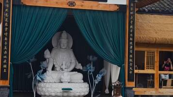 A Buddha statue in a temple entrance with green curtains and a traditional thatched roof structure.