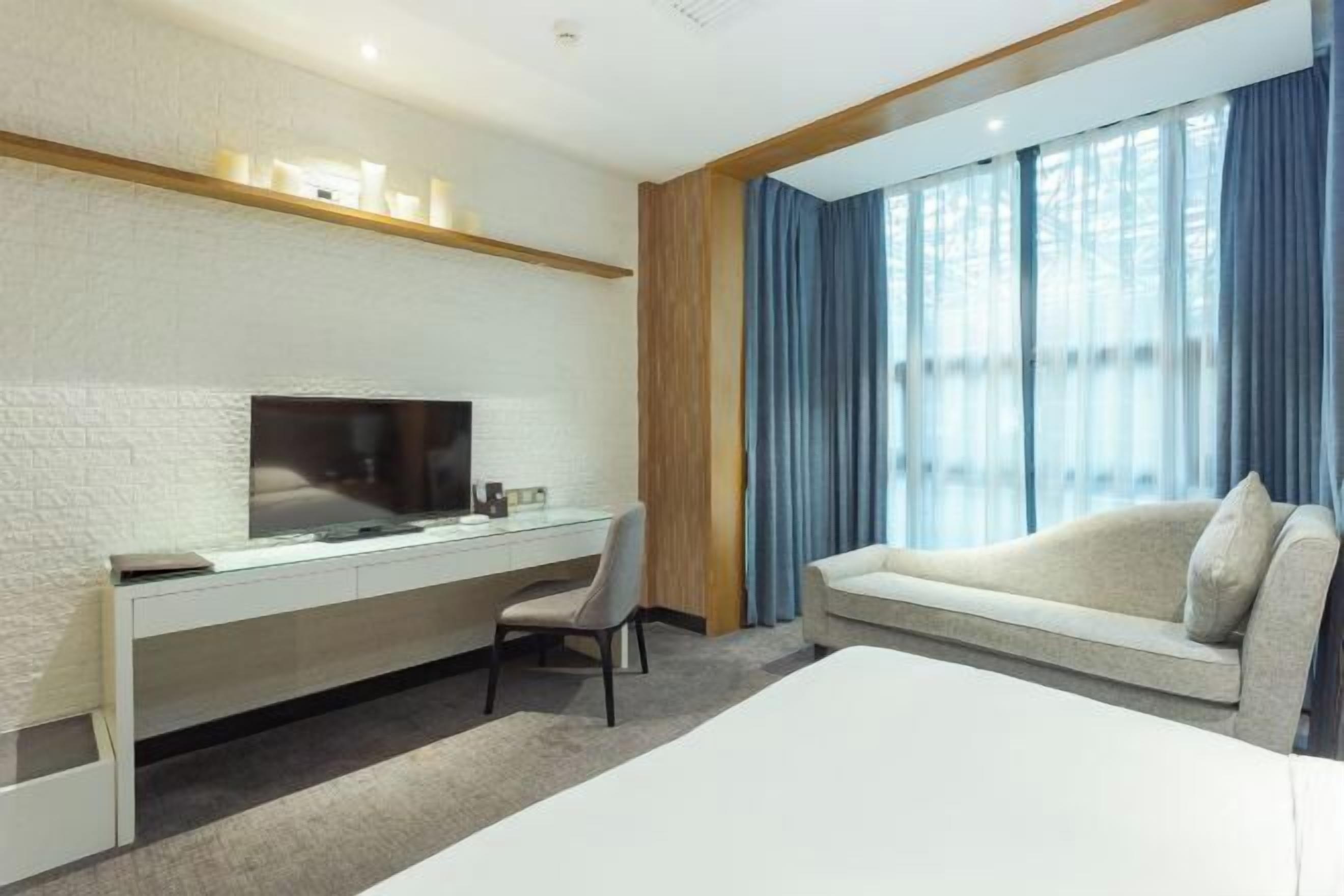 A modern hotel room with a flat-screen TV mounted on a white brick wall, a glass desk with a chair, a large bed, and a sofa.