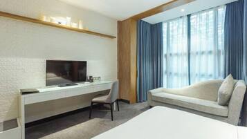 A modern hotel room with a flat-screen TV mounted on a white brick wall, a glass desk with a chair, a large bed, and a sofa.