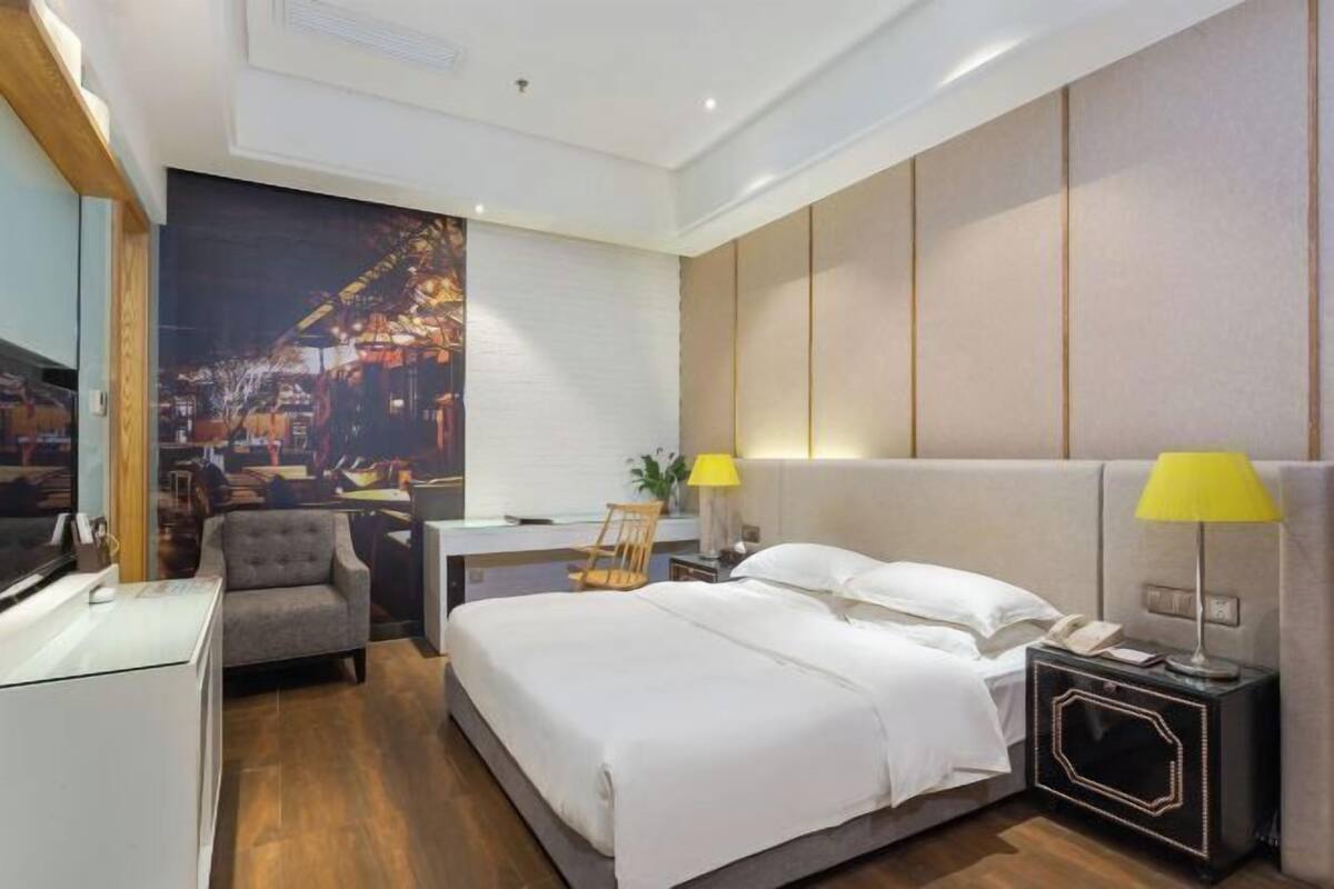 Une chambre d’hôtel comprenant un lit, une table de chevet, une lampe, une chaise, un bureau et un grand tableau représentant un paysage urbain accroché au mur.