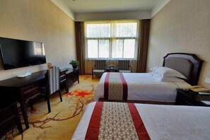 Quarto de hotel com duas camas, uma escrivaninha, televisão, uma cadeira e janela com cortinas.