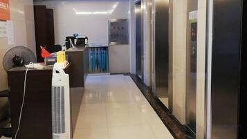 A hotel corridor with an elevator, reception desk, and a fan.