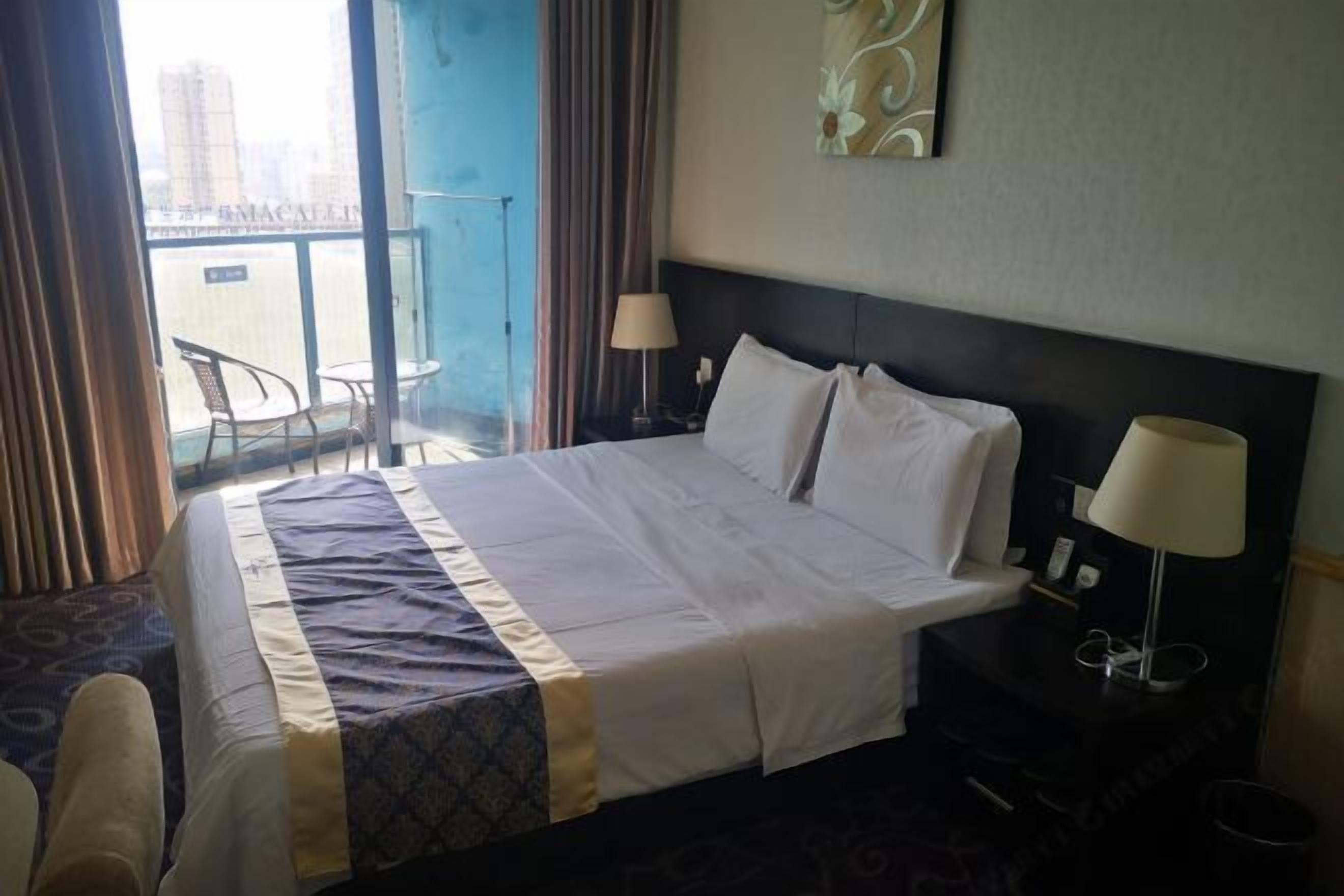 A hotel room with a large bed, two bedside lamps, a view of the city through the window, and a balcony with chairs.