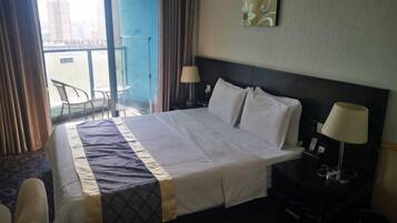 A hotel room with a large bed, two bedside lamps, a view of the city through the window, and a balcony with chairs.