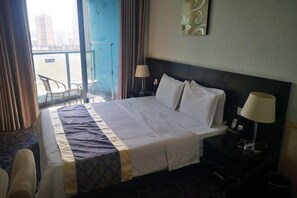 Habitación de hotel con una cama grande, dos lámparas de noche, vistas de la ciudad por la ventana y un balcón con sillas.