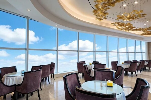 A spacious dining area with round tables and chairs, large windows offering a view of the sky.