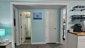A room with teal walls, a wooden floor, a white door, a kitchen area with a coffee maker, and a shelf with cups. - "Salty Dogs: Your Dog-Friendly Haven for Coastal Getaways!" (Oak Island)