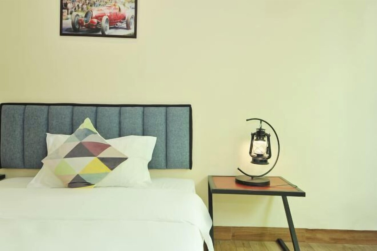 Una cama bien hecha con un almohadón de estampado geométrico, un cabecero, una mesita con un farol y un cuadro en la pared.