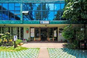 Un edificio con fachada de vidrio, un letrero que indica un lugar público y un acceso pavimentado con vegetación.