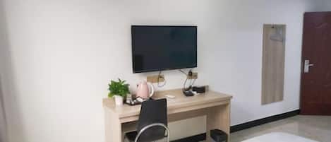 A hotel room with a bed, a desk with a chair, a television, a small plant, and a door.
