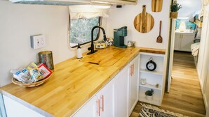 Een kleine keuken met een houten aanrecht, witte kastjes en een raam met zonwering.
