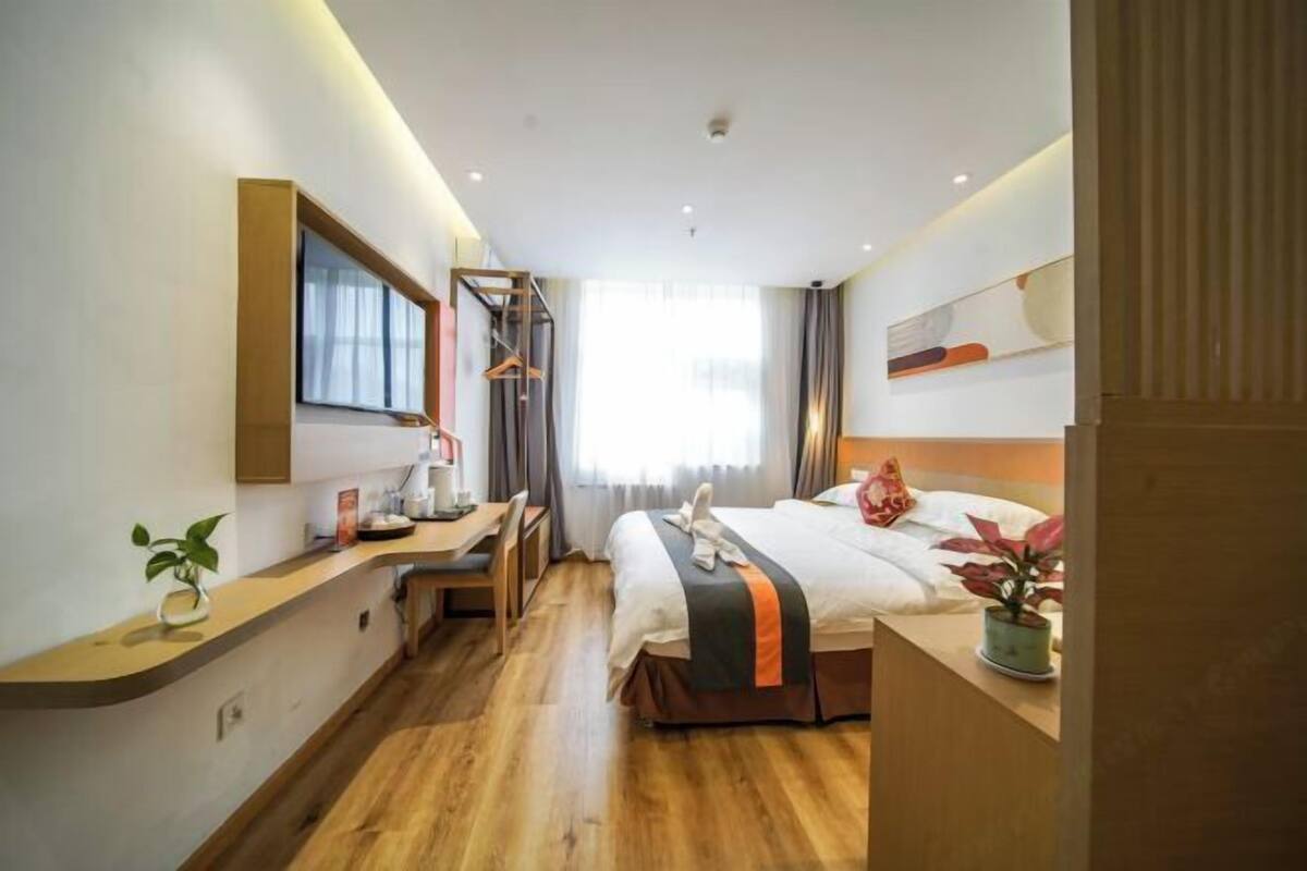 Habitació d'hotel moderna amb sòl de fusta, un llit amb edredó blanc i taronja, escriptori amb cadira, una petita planta i una finestra amb cortines.