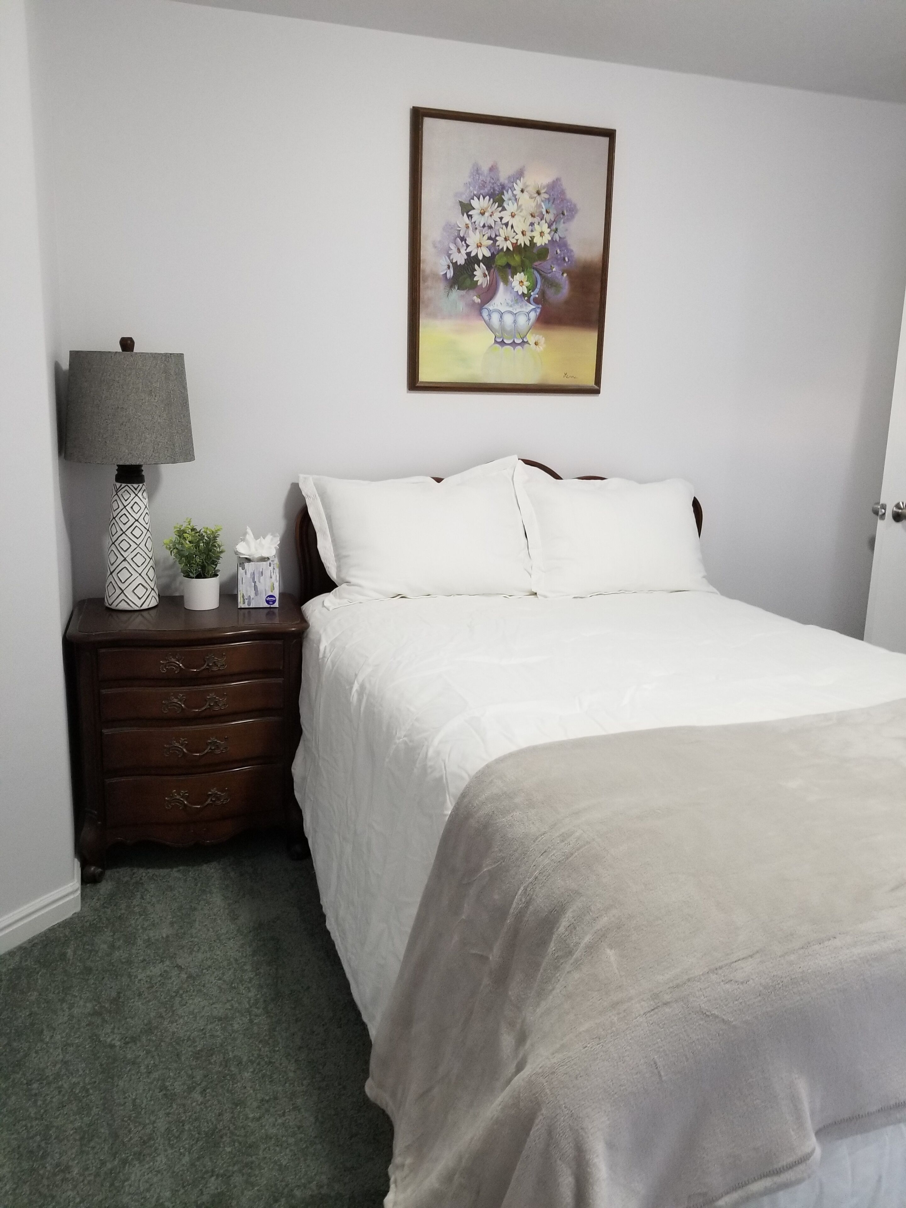 Een netjes opgemaakte slaapkamer met een bed, nachtkastje met een lamp, een kleine potplant en een bloemrijk schilderij aan de muur.