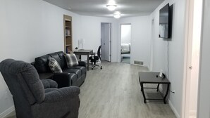 Un salón amplio con un sofá gris, una silla de escritorio negra, una mesita y un televisor montado en la pared.
