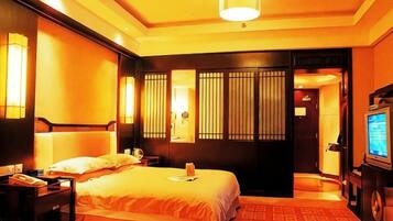 A hotel room with a bed, bedside table, lamp, TV, and a red carpet.