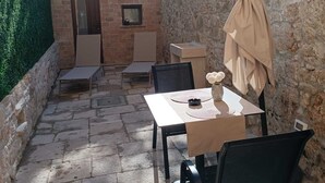 A patio area with stone flooring, a table with chairs, a small brick structure, and a green hedge.