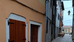 A narrow cobblestone street with buildings on either side, featuring a wooden door and shuttered window on the left.