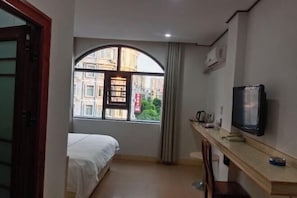 Ein kleiner, hell erleuchteter Raum mit einem Bett, einem Fernseher, einem Schreibtisch und einem Fenster mit Blick auf Gebäude.