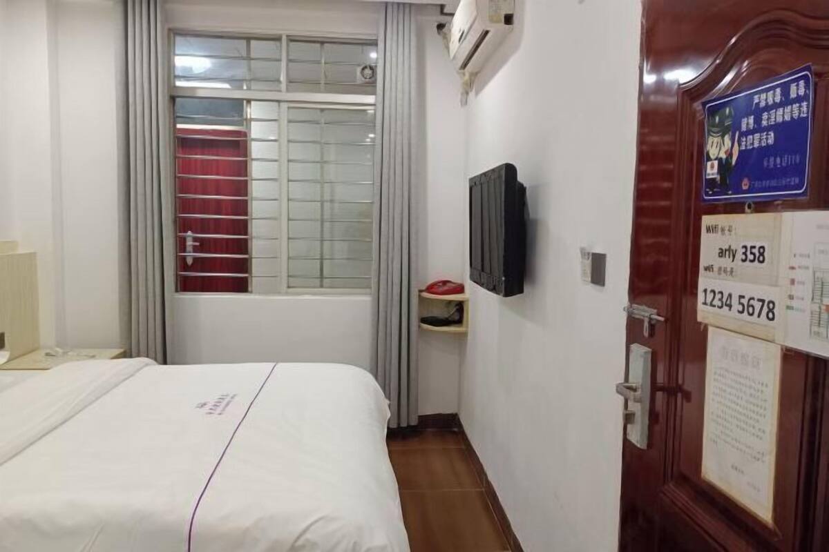 Une chambre d’hôtel avec un lit, un téléviseur fixé au mur, une fenêtre avec des rideaux et une porte portant un numéro.