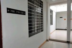 Un pasillo con una puerta identificada con los números 708 y 718-728, una ventana con celosía metálica y un suelo de baldosas.