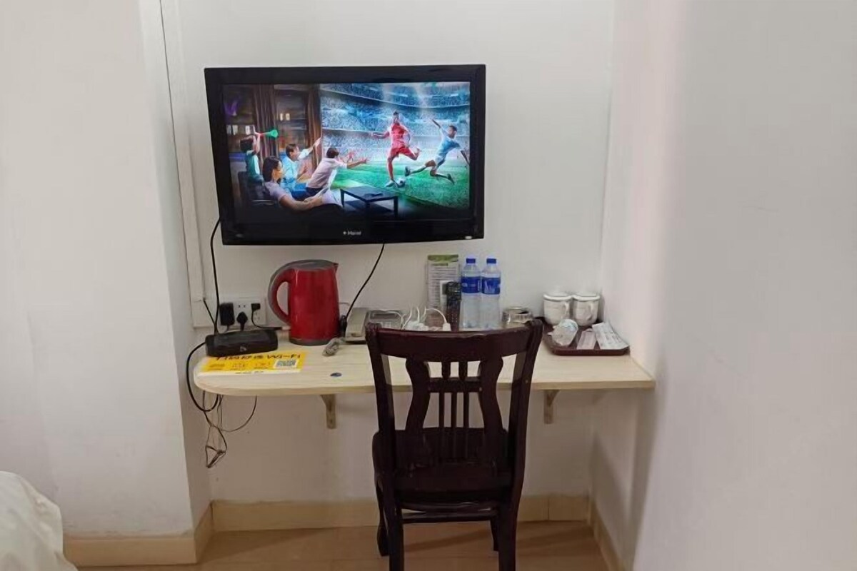 A television mounted on a wall displaying a sports event, a desk with a red mug, a white mug, and a bottle, and a wooden chair in front of the desk.