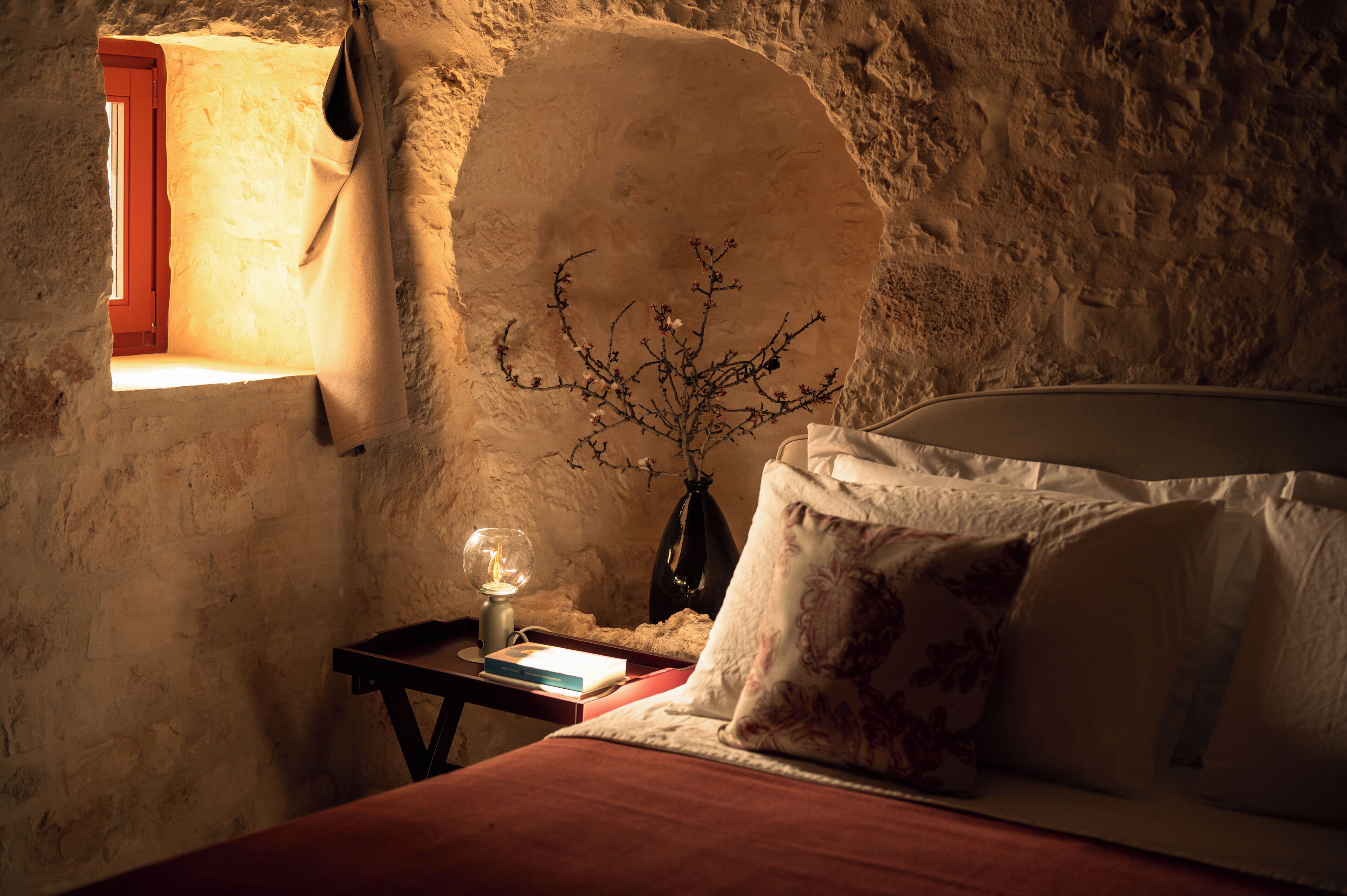 A bedroom with a stone wall, a window with a red frame, a bedside table with a lamp, and a bed with white bedding.