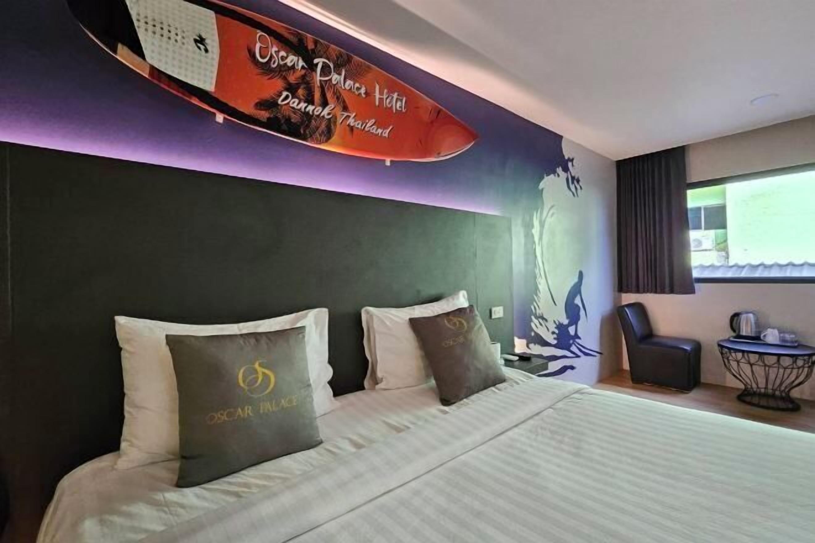 A hotel room with a bed, pillows, a chair, a small table, and a surfboard decoration.