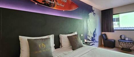 A hotel room with a bed, pillows, a chair, a small table, and a surfboard decoration.
