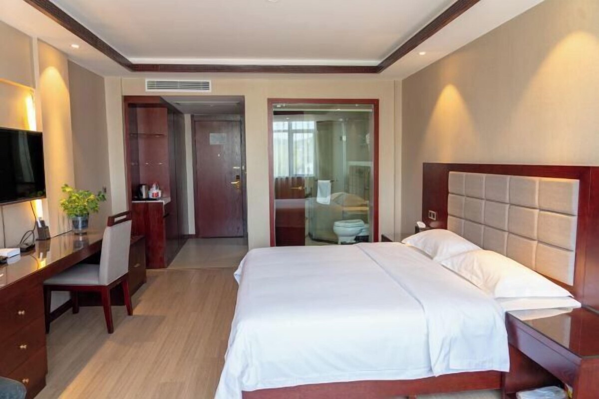 Une chambre d’hôtel équipée d’un lit, d’un bureau, d’une chaise, d’une télévision et d’une salle de bain visible à travers une porte en verre.