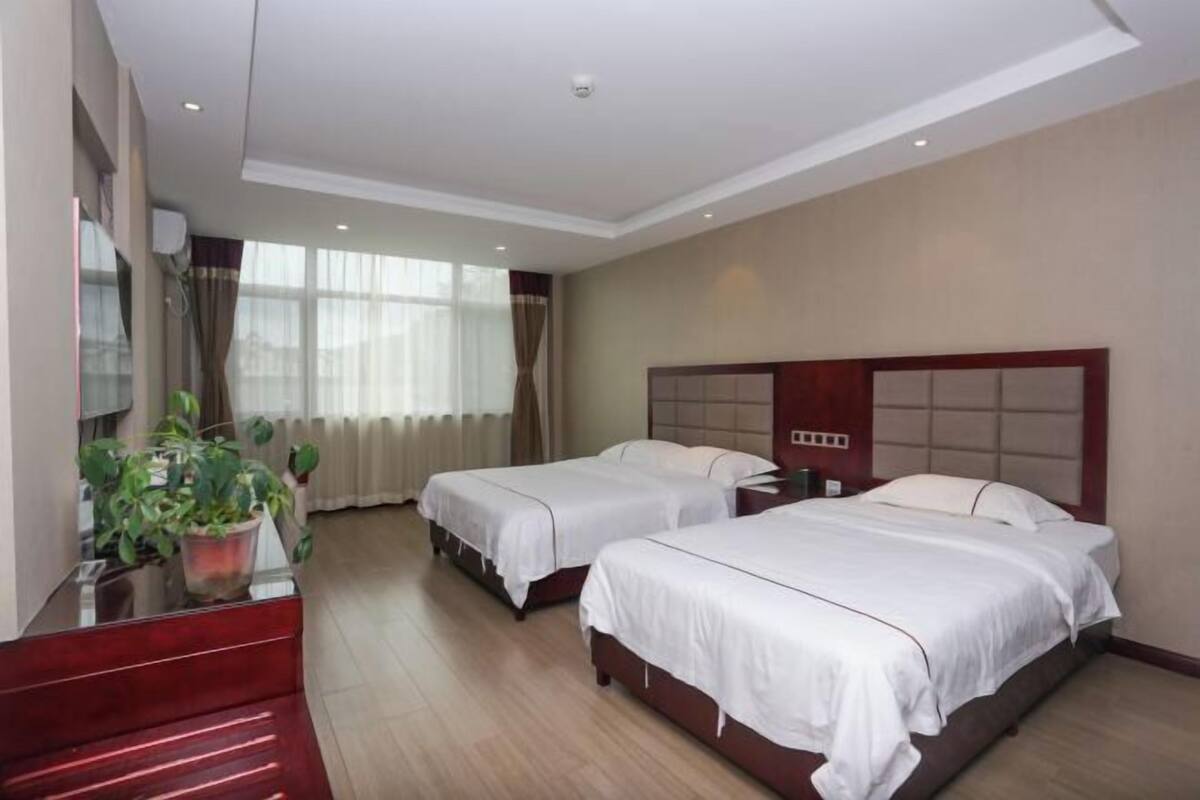 A hotel room with two beds, a large window with curtains, a potted plant, and a red bench.