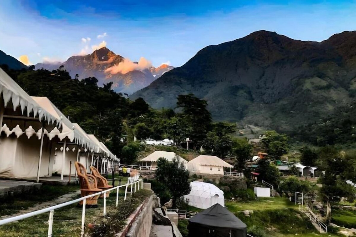 A mountainous landscape with a white building featuring a zigzag roof design, surrounded by greenery and other structures.
