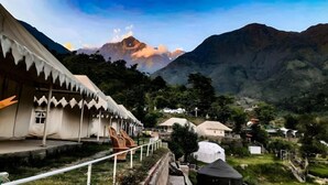 A mountainous landscape with a white building featuring a zigzag roof design, surrounded by greenery and other structures. - Hill Ventures (Dharamshala)