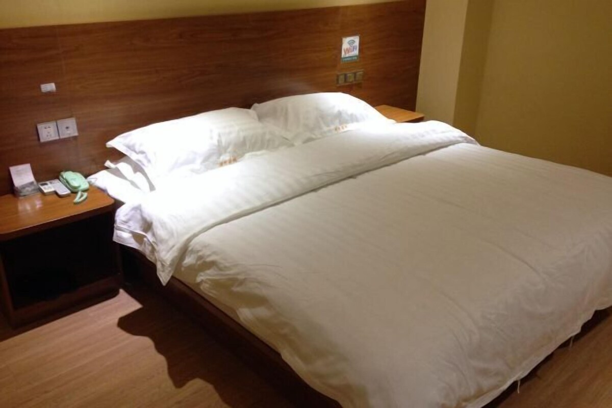 A hotel room with a wooden headboard, a bed with white linens, a bedside table with a phone and a lamp, and a wall-mounted power outlet.