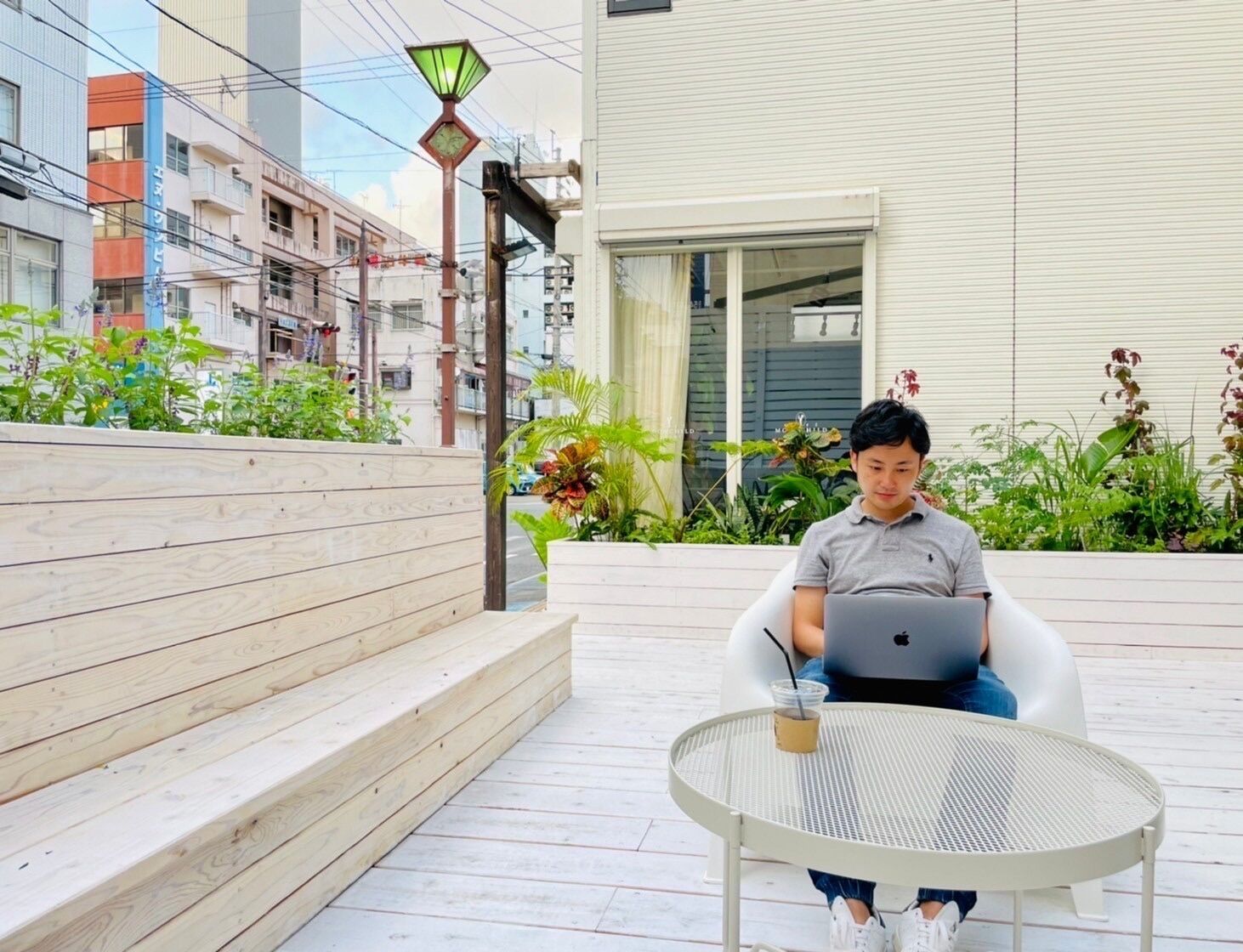 Um homem sentado numa cadeira branca com um laptop sobre uma mesa redonda de metal, ao ar livre, com um banco de madeira e plantas ao fundo.