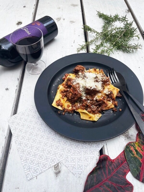 Une assiette de pâtes avec une sauce à la viande et du fromage râpé, un verre de vin rouge et une bouteille de vin sur une table en bois.