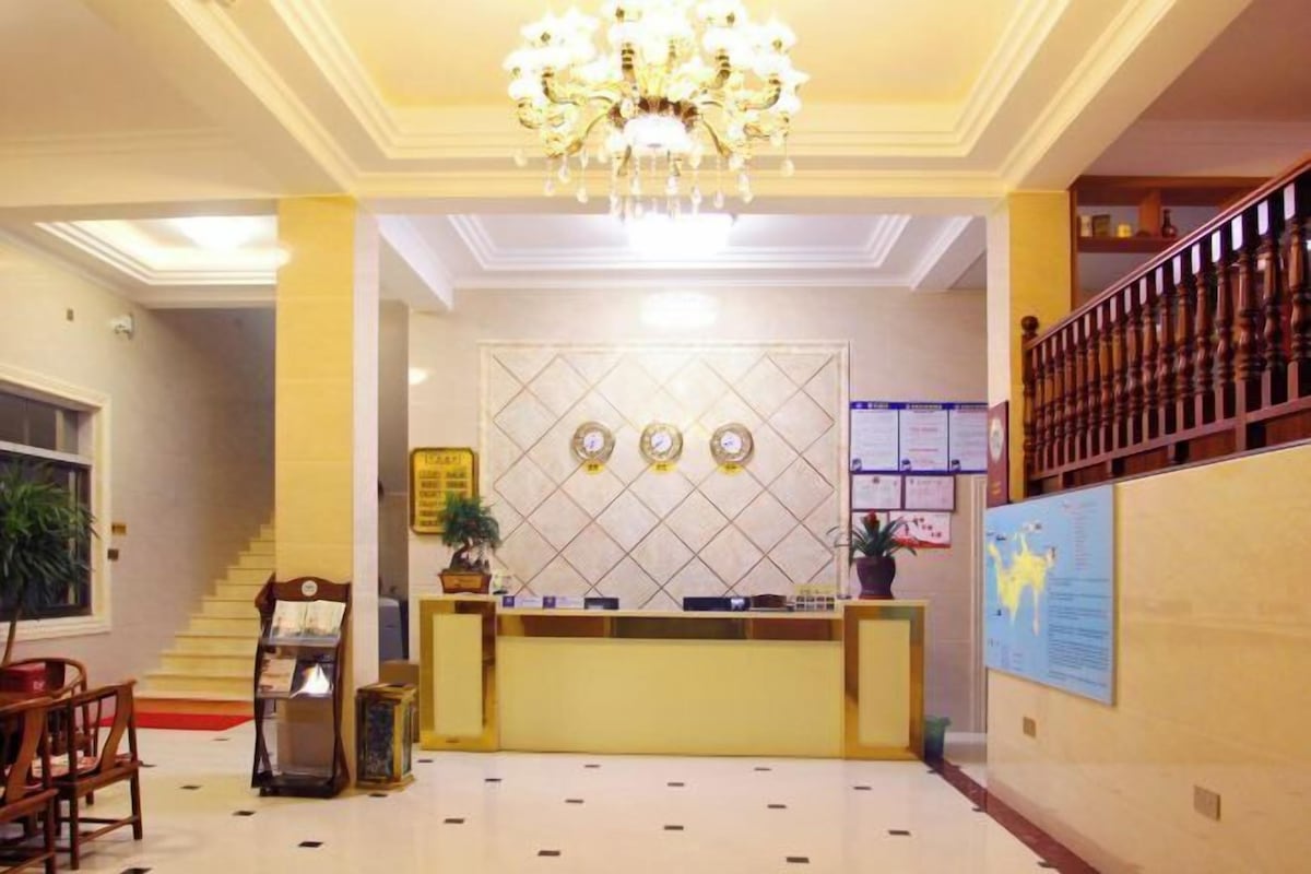 A hotel reception area with a chandelier, a reception desk, a staircase, and a wall with informational posters.