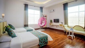 Une chambre d’hôtel avec deux lits, un coin salon comprenant un canapé et un fauteuil, un fauteuil suspendu rose et de grandes fenêtres avec des rideaux.