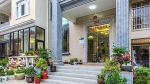 L’entrée d’un hôtel, avec un escalier menant à la porte, flanquée de plantes en pot et dotée d’une façade en verre transparent.