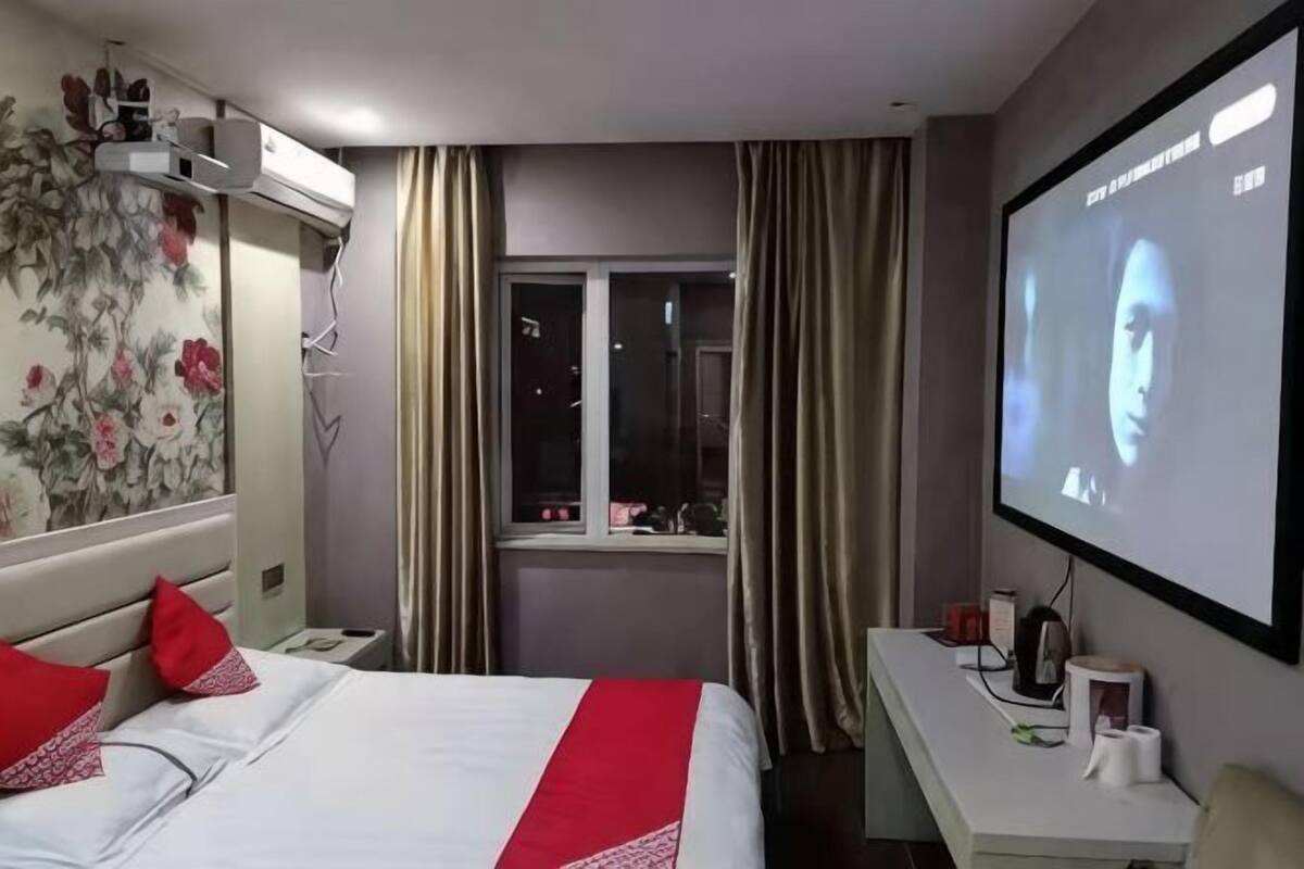 A hotel room with a bed, a TV screen, a desk with a computer, a window with curtains, and a floral wall decoration.