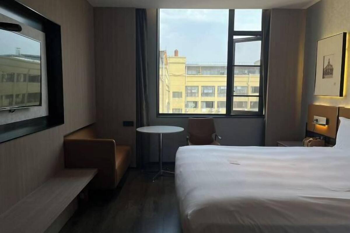 A hotel room with a bed, a small round table, a chair, a bench, and a large window with a view of buildings.