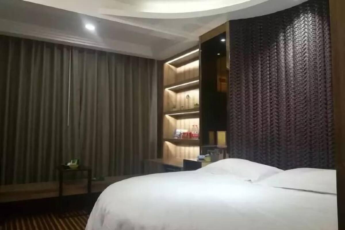 A hotel room with a large bed, a small table with a plant, and a built-in shelving unit with books and decorative items.