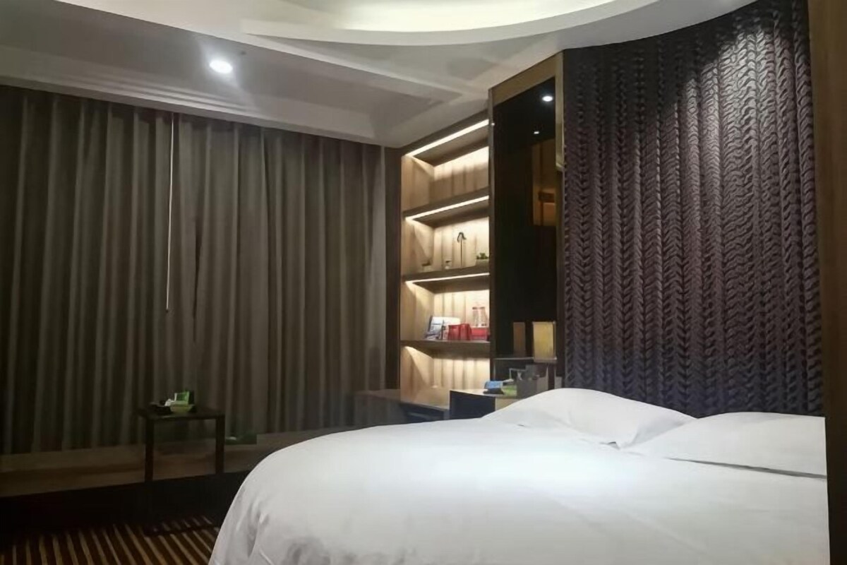 A hotel room with a large bed, a bedside table, a small table with a lamp, and a built-in shelf unit with lighting.