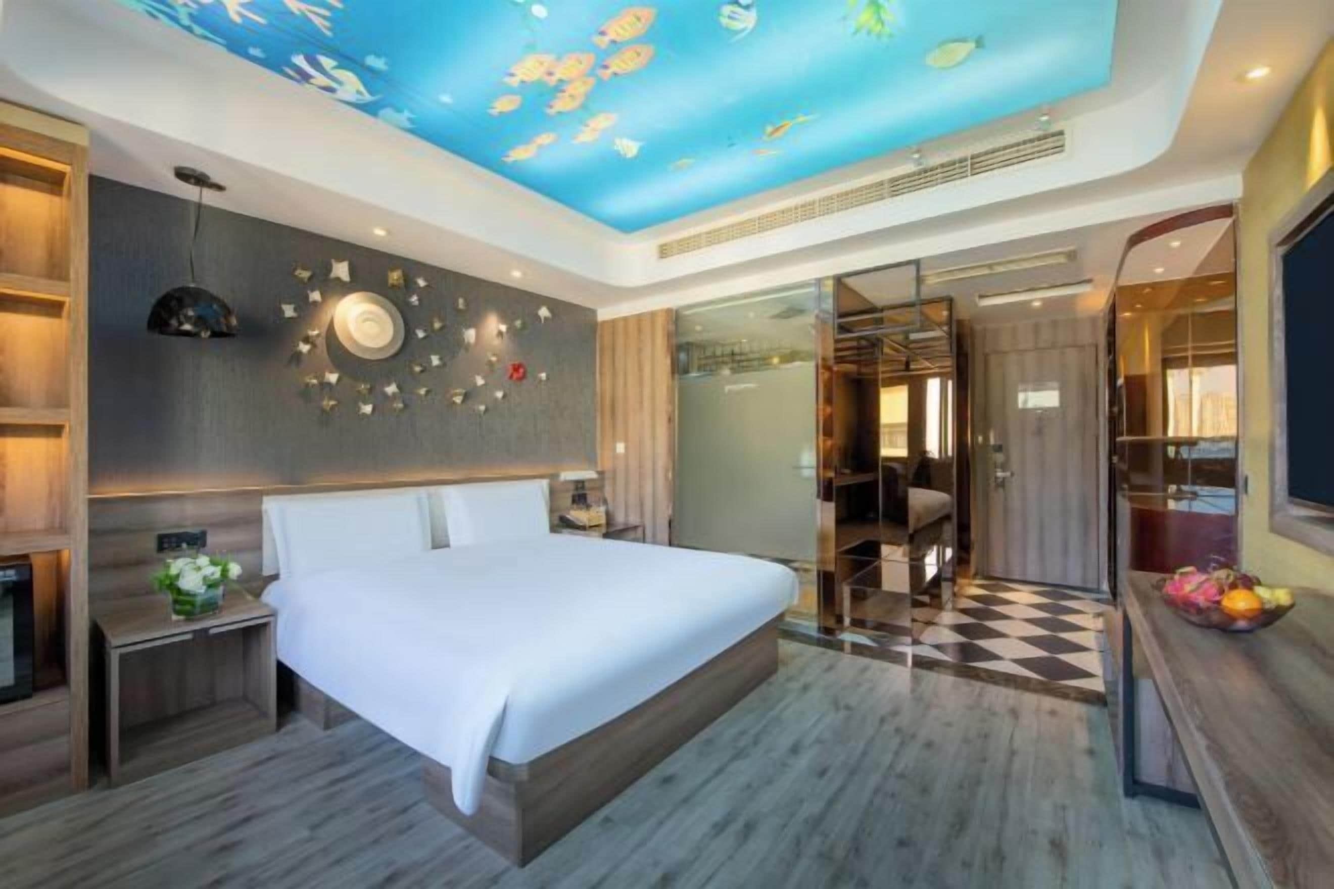 Habitación de hotel moderna con una cama grande, un mural en el techo con peces, un box de ducha de vidrio y piso a cuadros.