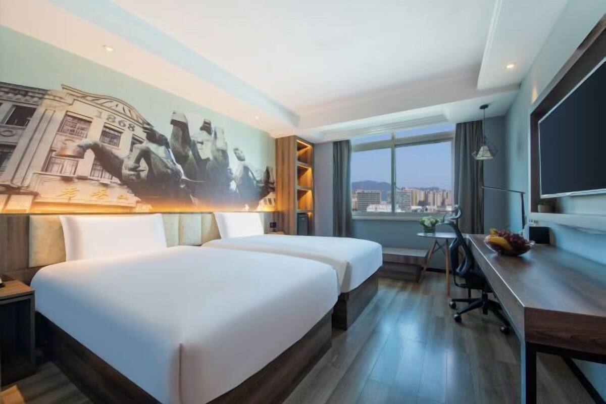 A hotel room with two beds, a desk, a chair, a TV, and a large window offering a city view.