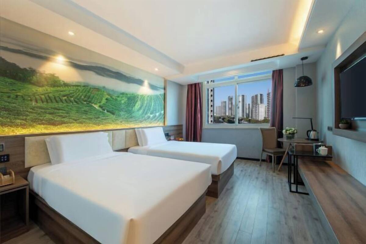 A hotel room with two beds, a desk, a chair, a window with a city view, and a painting on the wall.
