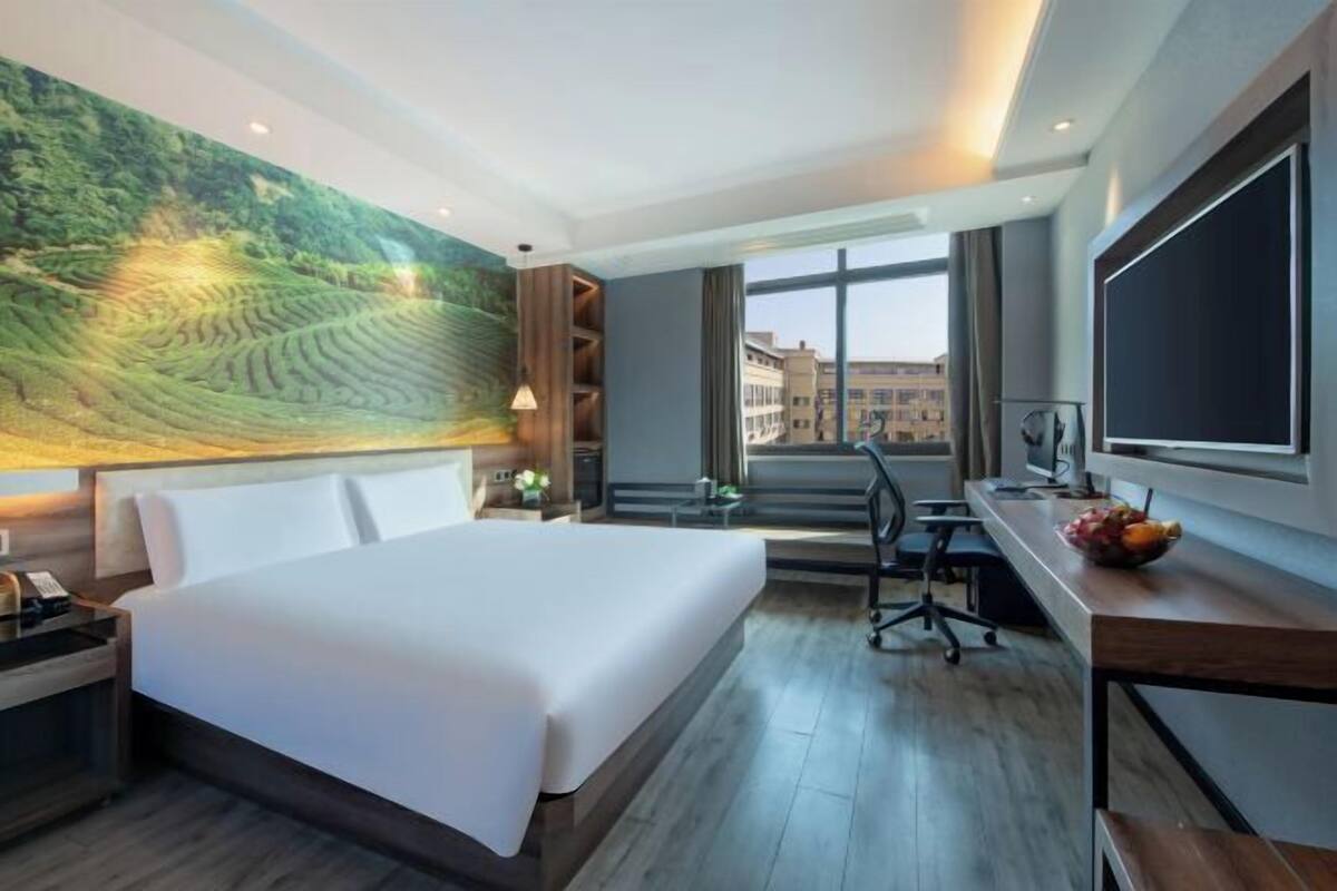 A hotel room with a large bed, a desk with a computer, a TV, and a large window with a city view.