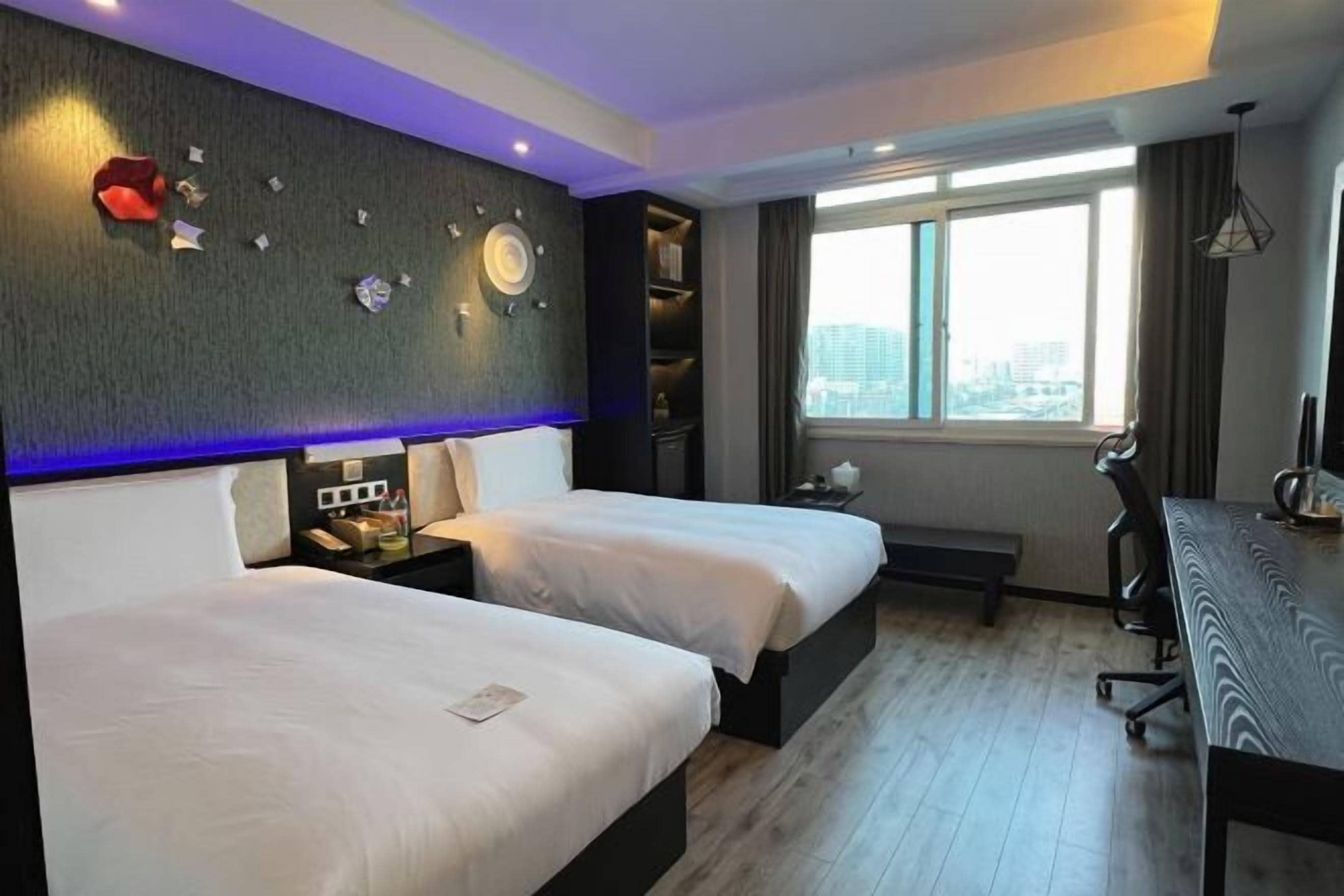 A hotel room with two beds, a desk, a chair, a bookshelf, and a window with a city view.